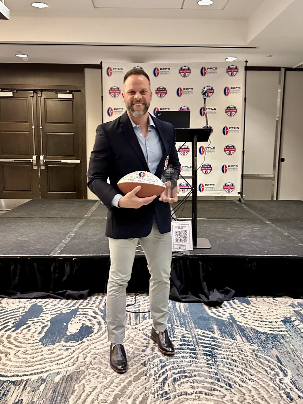 Dr. Joseph Krzemien holding the Professional Football Chiropractic Society Hall of Fame crystal trophy and signed induction football, on the PFCS Hall of Fame stage, February 28, 2025
